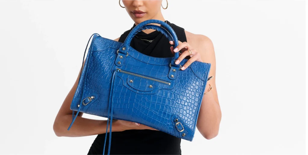 30 a model holds a bright blue crocodile embossed leather handbag with silver tone hardware against a plain white background